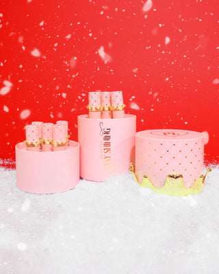 Set of pink cylindrical containers with gold accents containing 10 crown lip oils on a snowy background
