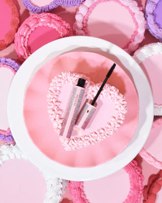 A tube of P.Louise Cry Baby Mascara with an applicator wand, displayed on top of a pink love heart cake surrounded by other pink cakes