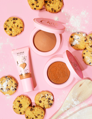 A collection of bronzers from P. Louise's Bake Me Bronzed Bronzer set, including a liquid bronzer, powder bronzer, and cream bronzer, presented with cookies and a makeup brush on a pink background.