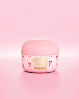 Pink tub of SLick It To Me shimmer melting body balm with decorative red bow elements on a pink background