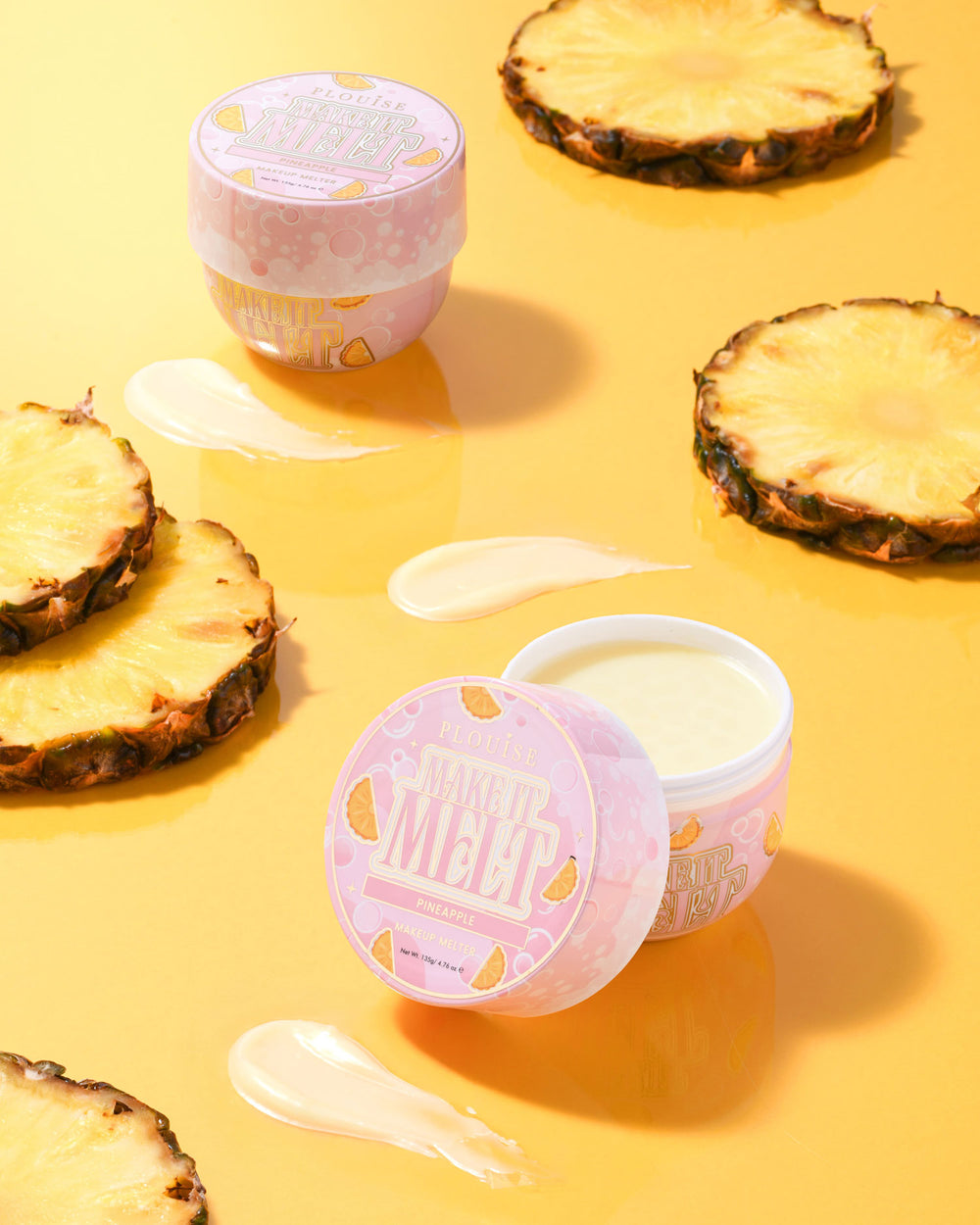 Twee kleine potjes P. Louise Make It Melt Makeup Removal Cleansing Balm in ananasgeur met een roze deksel, omgeven door ananasschijfjes op een gele achtergrond.