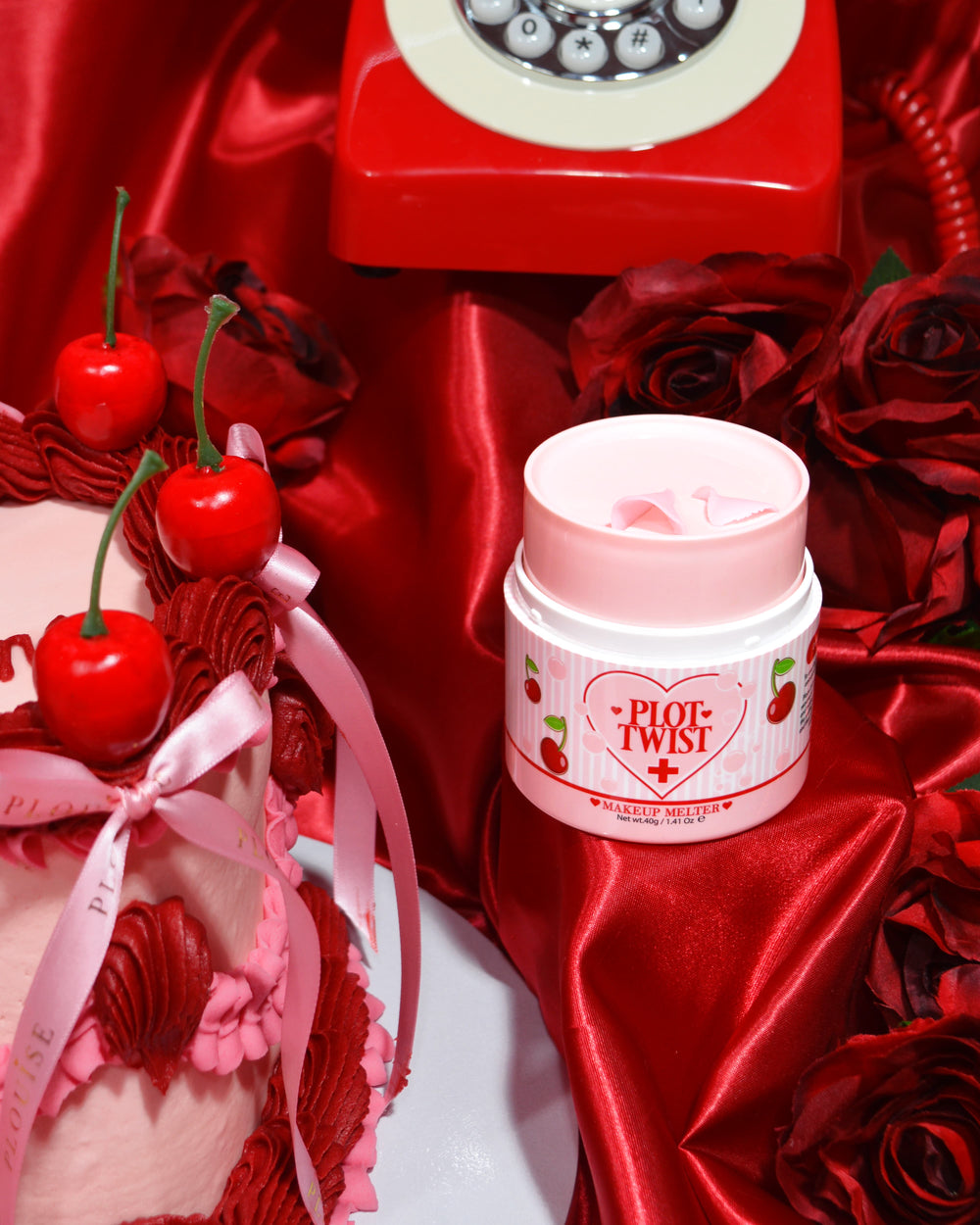 Een container met P.Louise Plot Twist Make It Melt Makeup Cleansing Balm weergegeven op een rood stoffen oppervlak met decoratieve elementen, waaronder een kers en een vintage rode telefoon op de achtergrond.