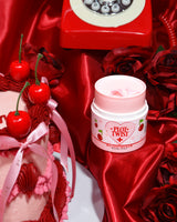 Een container met P.Louise Plot Twist Make It Melt Makeup Cleansing Balm weergegeven op een rood stoffen oppervlak met decoratieve elementen, waaronder een kers en een vintage rode telefoon op de achtergrond.