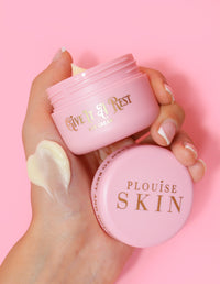 Een persoon die een roze pot P.Louise Give It A Rest oogcrème vasthoudt met de deksel eraf, met de productnaam en het merk op de pot.