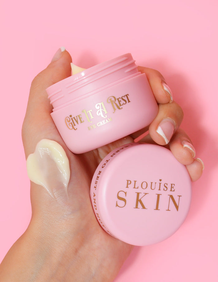 Een persoon die een roze pot P.Louise Give It A Rest oogcrème vasthoudt met de deksel eraf, met de productnaam en het merk op de pot.